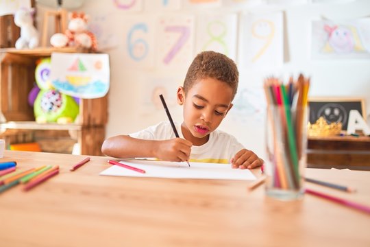 Beautiful african american toddler sitting drawing using paper and pencils on desk at kindergarten
