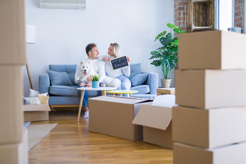 Obraz premium Young beautiful couple with dog sitting on the sofa holding blackboard with message at new home around cardboard boxes