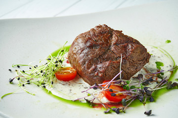 Delicious medium-rare beef steak with a side dish of microgreen and cherry tomatoes on a white plate on a white wooden background. Close up. Food. Copy space