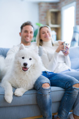Young beautiful couple with dog sitting on the sofa drinking coffee at new home around cardboard boxes