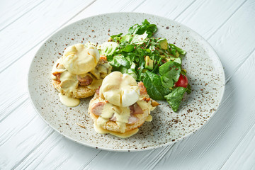 Breakfast with toasts with creamy garlic sauce, bacon and poached eggs with lettuce on a ceramic plate on a white wooden background