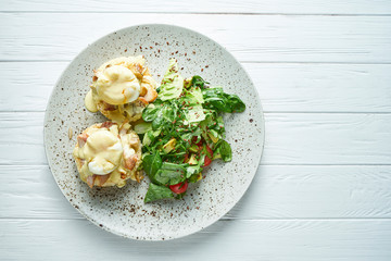 Breakfast with toasts with creamy garlic sauce, bacon and poached eggs with lettuce on a ceramic plate on a white wooden background
