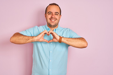 Young man wearing blue casual shirt standing over isolated pink background smiling in love showing heart symbol and shape with hands. Romantic concept.