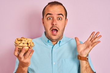 Young man holding bowl with peanuts standing over isolated pink background very happy and excited, winner expression celebrating victory screaming with big smile and raised hands