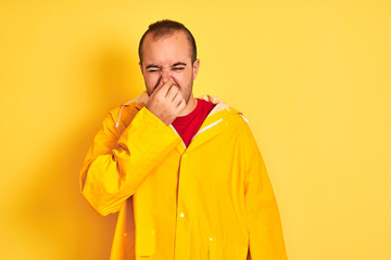 Young man wearing rain coat standing over isolated yellow background smelling something stinky and disgusting, intolerable smell, holding breath with fingers on nose. Bad smells concept.