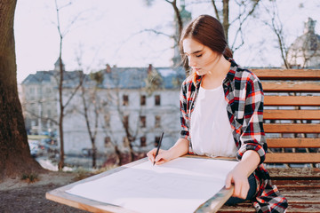 Young woman artist draws a picture while sitting on a bench. Painter artist holds a wooden tablet for painting and a brush in his hand