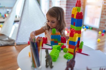 Adorable blonde toddler playing with building blocks around lots of toys at kindergarten