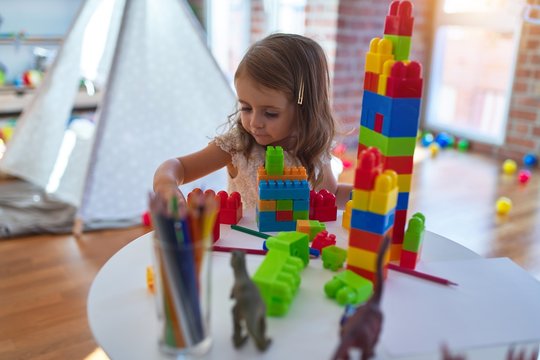 Adorable blonde toddler playing with building blocks around lots of toys at kindergarten