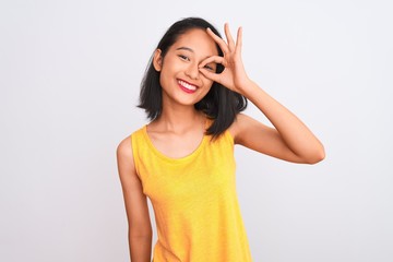 Young chinese woman wearing yellow casual t-shirt standing over isolated white background doing ok gesture with hand smiling, eye looking through fingers with happy face.