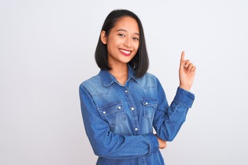 Young beautiful chinese woman wearing denim shirt standing over isolated white background with a big smile on face, pointing with hand and finger to the side looking at the camera.