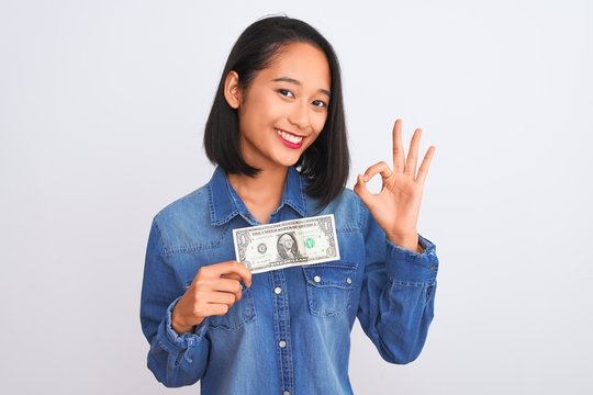 Young beautiful chinese woman holding one dollar standing over isolated white background doing ok sign with fingers, excellent symbol