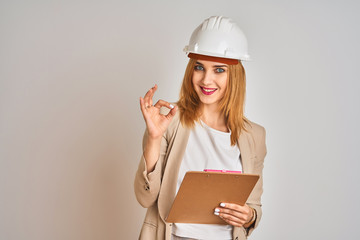 Redhead caucasian business woman wearing safety helmet and holding clipboard doing ok sign with fingers, excellent symbol