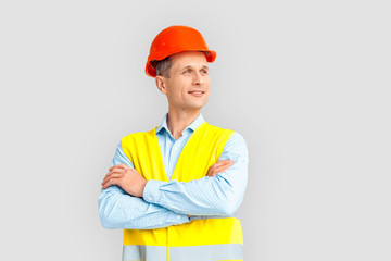 Construction. Mature man in hardhat and uniform standing isolated on white crossed arms looking aside dreamful