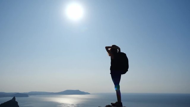 Sportswoman stands on a tall rock and enjoys a charming landscape, views of the sea and the blinding rays of the sun in slow motion, back view. Healthy lifestyle and travel through the mountains.