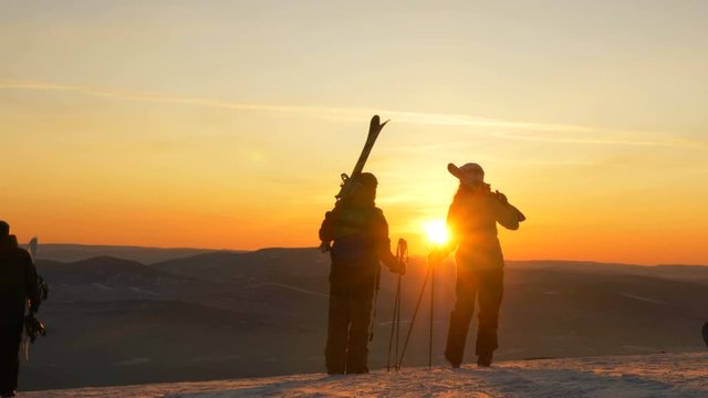 skiers with equipment stand on high hilltop admiring sunset