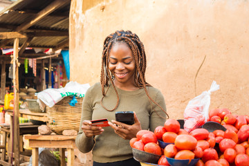 young african woman in a local african market using her mobile phone and credit card for a...