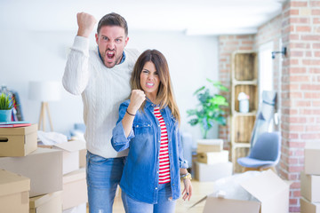 Young beautiful couple standing at new home around cardboard boxes angry and mad raising fist frustrated and furious while shouting with anger. Rage and aggressive concept.