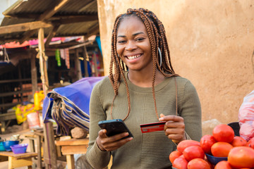 nigerian woman selling in a local nigerian market using her mobile phone and credit card to do a...