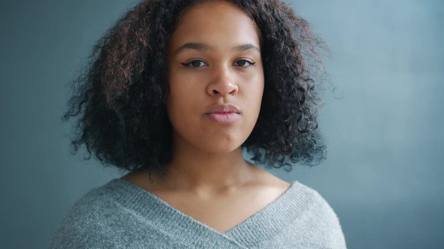 Portrait of attractive Afro-American girl looking at camera with serious face standing alone against dark gray background. Confident youth and emotions concept.