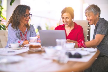 Meeting of middle age women having lunch and drinking coffee. Mature friends smiling happy using laptop at home on a sunny day