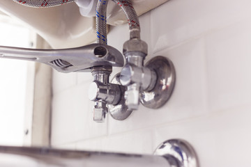 Plumber turns nut on angle valve under wash basin using spreading wrench.