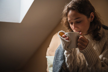 Beautiful young woman drinking coffee in a room with winter clothes, resting at home on a winter morning