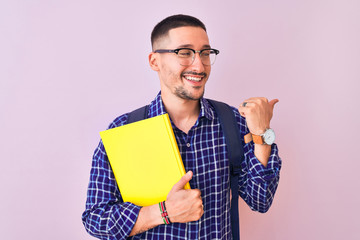 Young handsome student man holding a book over isolated background pointing and showing with thumb up to the side with happy face smiling