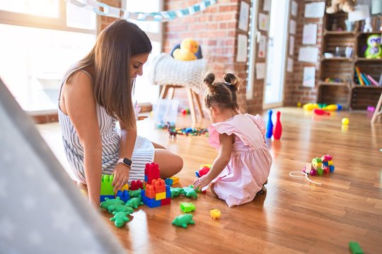 Young beautiful teacher and toddler sitting on the floor playing with building blocks toy at kindergarten