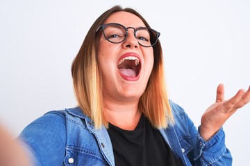 Beautiful woman wearing denim shirt and glasses make selfie over isolated white background very happy and excited, winner expression celebrating victory screaming with big smile and raised hands