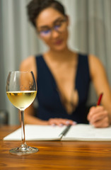 A woman wearing glasses sits at a table drinking white wine and writing in a journal