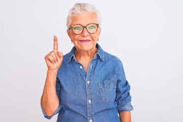 Senior grey-haired woman wearing denim shirt and glasses over isolated white background showing and pointing up with finger number one while smiling confident and happy.