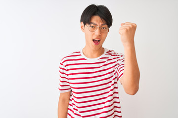 Young chinese man wearing glasses and striped t-shirt standing over isolated white background angry and mad raising fist frustrated and furious while shouting with anger. Rage and aggressive concept.