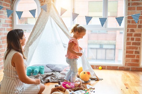Beautiful teacher and blond toddler girl playing with dolls inside tipi at kindergarten