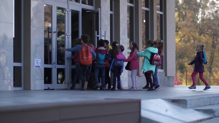 Side view of joyful elementary age schoolkids hurrying to enter school doors. Crowd of diverse little pupils with backpacks running while rushing to start lessons early morning