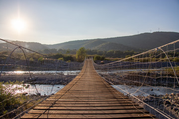 Puente colgante de madera sobre un rio lleno de rocas, cielo azul y plantas