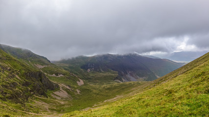 A scenic view of a mountain valley with grassy slopes, cliff, trail and mountain range in the background under a stormy grey cloudy sky