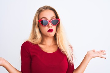 Young beautiful woman wearing red t-shirt and sunglasses over isolated white background clueless and confused expression with arms and hands raised. Doubt concept.