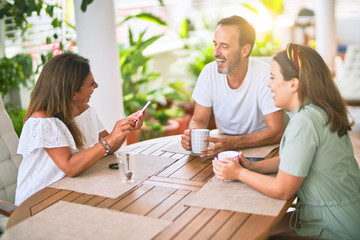 Beautiful family sitting on terrace drinking cup of coffee using smartphone speaking and smiling