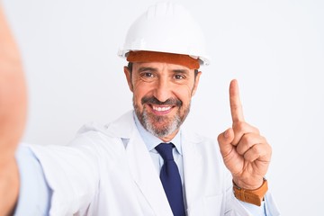 Middle age engineer man wearing helmet make selfie over isolated white background surprised with an idea or question pointing finger with happy face, number one