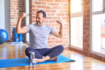 Middle age handsome sportman sitting on mat doing stretching yoga exercise at gym showing arms...