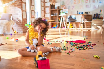 Beautiful toddler wearing glasses and unicorn diadem sitting on the floor at kindergarten thinking...