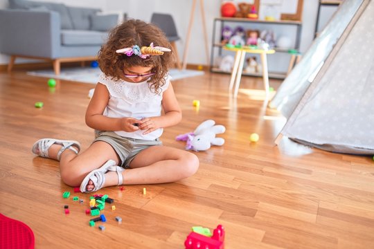 Beautiful toddler wearing glasses and unicorn diadem sitting on the floor playing with building blocks at kindergarten