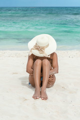 Fototapeta na wymiar Mujer sentada en la arena abrazando sus piernas. Mujer vistiendo traje de baño y sombrero en la playa. Mar al fondo. Mujer bronceada. Tomando el sol. Concepto de vacaciones y relajación