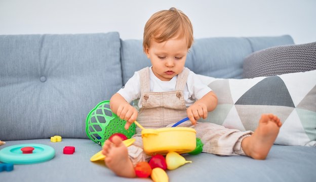 Adorable blonde toddler sitting on the sofa playing with plastic meals toys at home