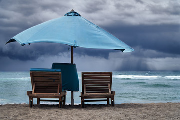 Storm on the sea with thunderclouds. Empty beach with umbrella and sun beds on background of hard rain over ocean.