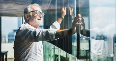 Retirement concept. Senior grey-haired businessman standing and looking to right hand at modern business lounge high up in an office tower.