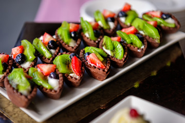 Delicious sweet tartlets made with chocolate dough with fresh fruit and berries close-up. Small cakes with strawberries, kiwi, grapes. Banquet service at wedding, anniversary.