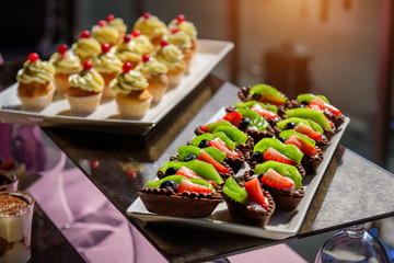 Cakes with fresh fruit and berries on the holiday table, close-up. Cupcakes with cream. Sweet table at gala dinner.