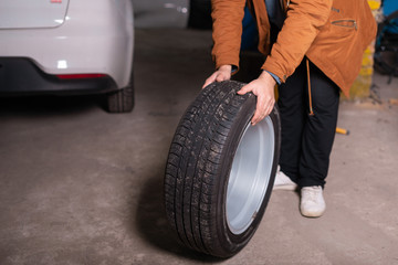 Obraz premium A man in an orange jacket holds a wheel against the background of a car, a hundred