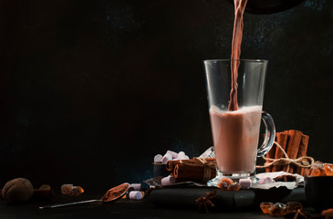 Pouring winter hot chocolate cocoa drink into tall glass with ispices on black table. Dark blue...